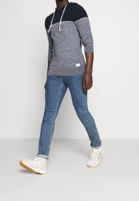 Mäns hooded tröja i mörk marinblå och grått stickat material, med snörning, långa ärmar, matchad med ljusblå skinny jeans och vita sneakers.