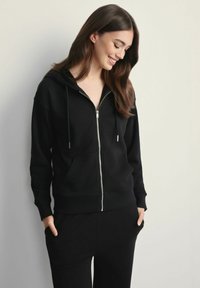 Młoda kobieta uśmiechnięta, ubrana w czarną bluzę z kapturem na zamek, z rękami w kieszeniach, stojąca na tle gładkiej jasnej ściany.