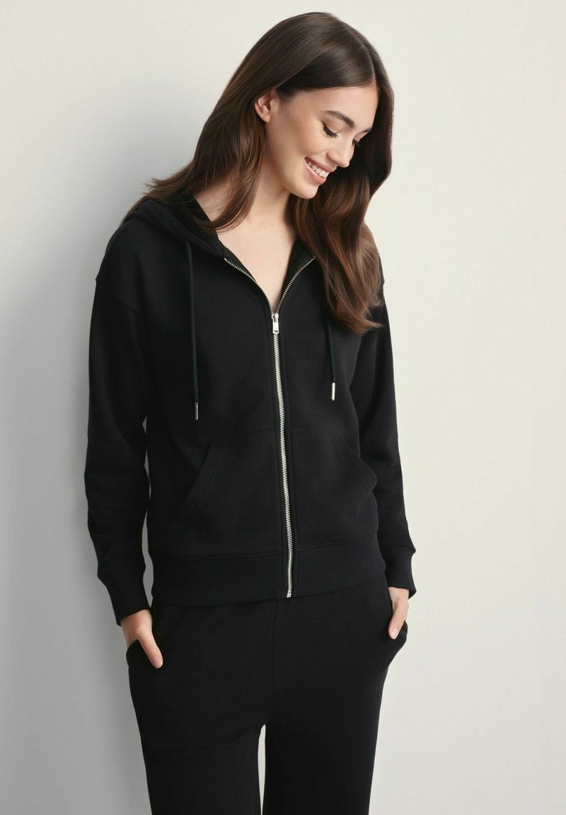 Młoda kobieta uśmiechnięta, ubrana w czarną bluzę z kapturem na zamek, z rękami w kieszeniach, stojąca na tle gładkiej jasnej ściany.