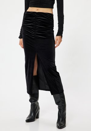 Zwarte fluwelen midi-rok met geplooide details, een zijsplit en een zachte textuur. Draagt met kniehoog zwarte laarzen met hak.