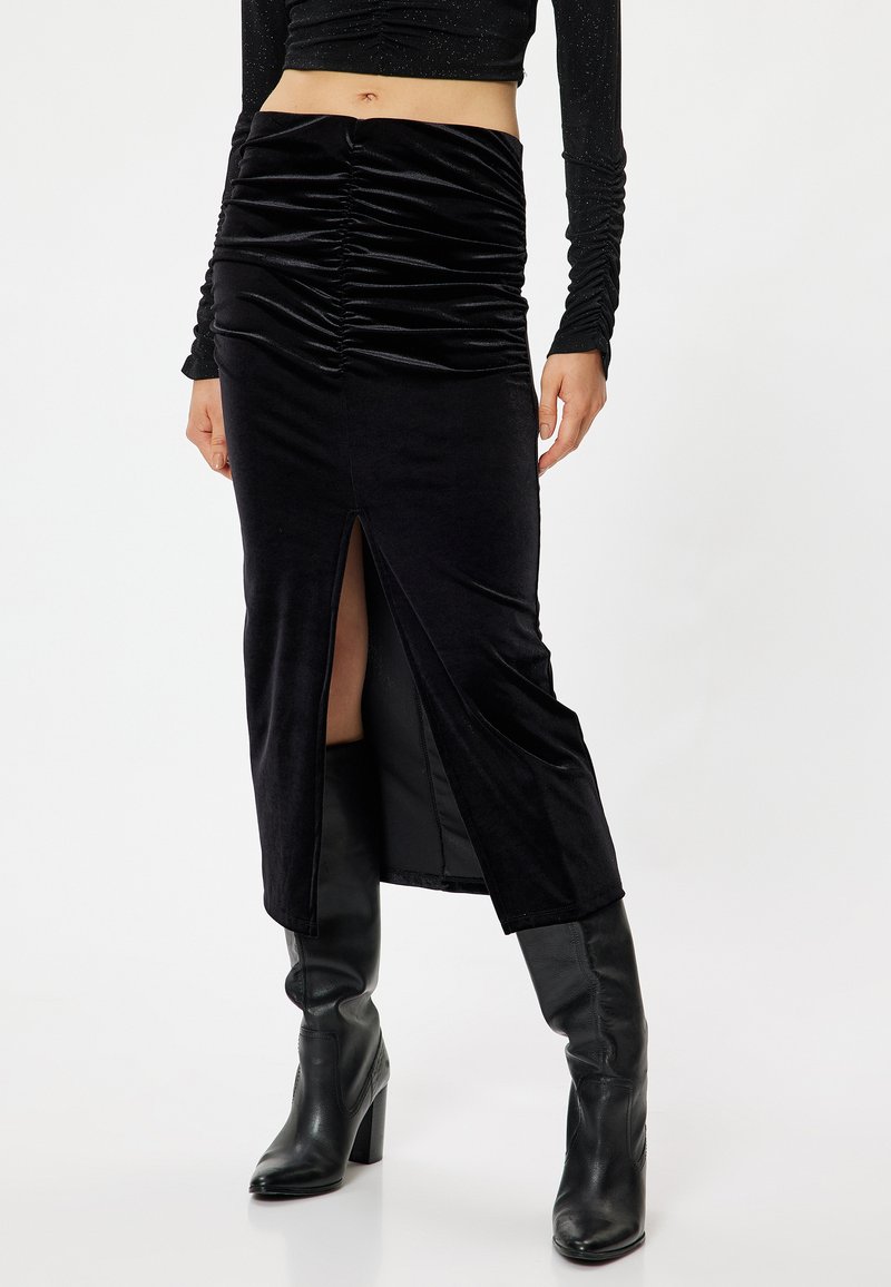 Zwarte fluwelen midi-rok met geplooide details, een zijsplit en een zachte textuur. Draagt met kniehoog zwarte laarzen met hak.
