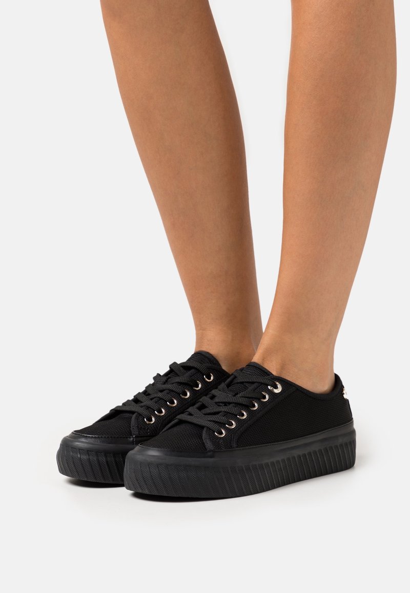 Schwarze Sneakers mit niedrigem Schnitt, dicken, gerippten Sohlen und schwarzen Schnürsenkeln, die auf baren Füßen vor weißem Hintergrund getragen werden.