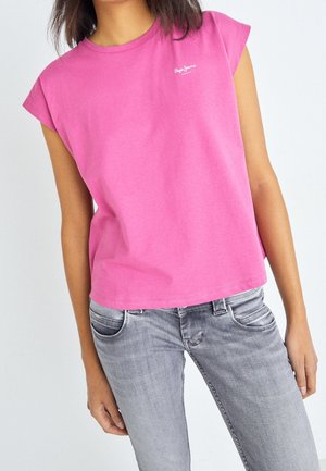 Personne portant un haut rose vif sans manches avec un petit logo blanc et un jean gris délavé, se tenant devant un fond blanc uni.