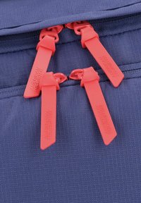 Primo piano di uno zaino blu navy con quattro tirazip rossi vivaci contrassegnati con un marchio in rilievo, che mette in mostra una superficie in tessuto testurizzato.