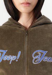 Maslinasto zelena zip-up dukserica s mekom teksturom. Ima plavi izvezeni natpis "Joop! Jeans" sprijeda i kapuljaču. Detalj sa zatvaračem je vidljiv.
