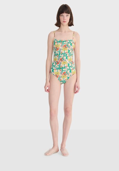 Jeune femme debout, portant un maillot de bain une pièce avec un motif floral et d'oiseaux vert, jaune et orange, ainsi que des chaussures blanches à enfiler en mesh.