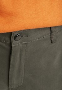 Bouton noir fermant un pantalon vert olive porté avec un pull en tricot orange, montré en gros plan à la taille.