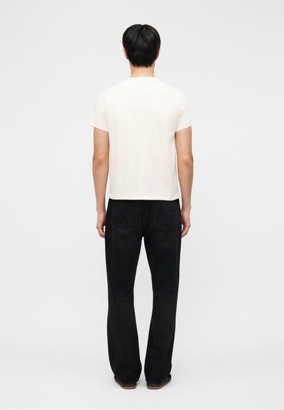 Homme debout, de dos, portant un t-shirt blanc uni à manches courtes et un jean noir droit sur un fond gris clair.