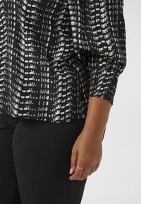 Sort bluse med et struktureret hvidt geometrisk mønster, samlede ærmer og subtil elasticitet ved manchetterne, parret med mørk denim.