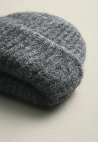 Beanie grigio in lana con una superficie texturizzata, caratterizzato da un design morbido a maglia e una visiera ripiegata. Il tessuto appare soffice e caldo.