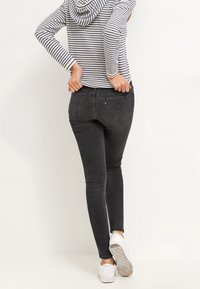 Persona che indossa jeans neri e sneakers bianche, mentre sistema le tasche posteriori, vestita con una maglia a maniche lunghe a righe, in piedi contro un muro bianco.