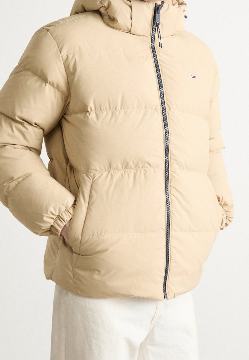 Homme portant une doudoune beige avec fermeture éclair à l'avant, poignets élastiques, poches latérales, et petit logo sur le côté gauche de la poitrine, portée avec un pantalon blanc.