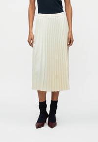 Fustă midi pliată în crem, cu o textură fină și o talie ajustată. Asortată cu șosete în dungi închise și pantofi cu vârf ascuțit.