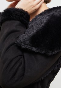Černý kabát s měkkým límcem z umělé kožešiny, strukturovaný tvar, texturovaná tkanina a na ramenou řasení pro přidanou dimenzi.
