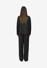 Personne avec de longs cheveux bruns portant un haut noir à manches longues et un pantalon noir ample, se tenant de dos sur un fond blanc.