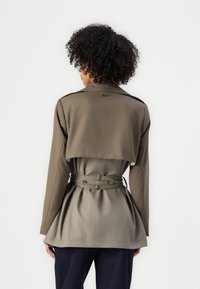 Oliven grønn omslagsjakke med kort design, lange ermer, belte i midjen og plissert bak. Har krage og to knapper.