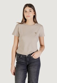 T-shirt di cotone beige con scollatura rotonda, maniche corte e un piccolo logo a forma di triangolo sul lato sinistro del petto. Indossato con jeans scuri.