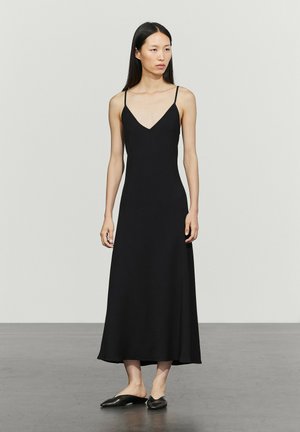 Frau mit langen schwarzen Haaren, die ein ärmelloses schwarzes Midikleid und schwarze Slip-On-Schuhe trägt, steht auf einem grauen Boden vor einer weißen Wand.