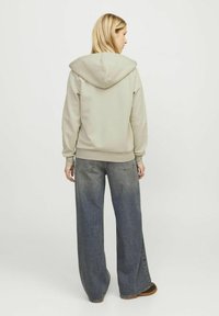 Haut à capuche gris clair avec un cordon, associé à un jean en denim foncé à jambe large. Texture lisse et coupe décontractée, avec des détails de couture visibles.