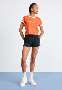 T-shirt à manches courtes rouge-orangée avec un col et des poignets beige, shorts noirs avec cordon de serrage, et baskets blanches avec des semelles texturées et des chaussettes légères.
