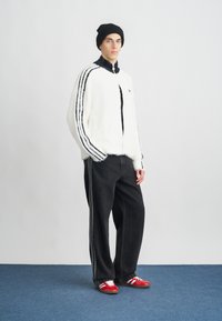 Chaqueta blanca y esponjosa con cierre, con rayas negras, combinada con jeans anchos negros y zapatillas rojas con rayas blancas. Se lleva un gorro negro.