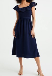 Marineblauwe jurk met off-shoulder oogjes ruffles, geplooide taille en vloeiende midi-lengte rok. Zachte, gestructureerde stof. Platte schoenen.