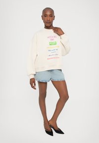 Krémová mikina s viacfarebným nápisom "Moschino Jeans", skombinovaná s svetlými denimovými šortkami a čiernymi špicatými balerínkami.