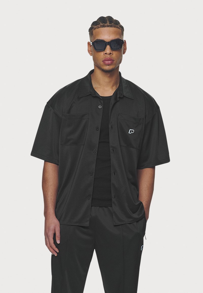 Black short-sleeve button-up shirt with two chest pockets, made from a smooth fabric, paired with black pants, featuring a small logo.