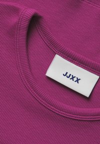 Fuksijos spalvos raukšlėtas audinys, suapvalintu kaklu, su baltu etiketė, ant kurios rašoma "JJXX" tamsiai mėlyna spalva. Artimas apykaklės ir etiketės detalės vaizdas.