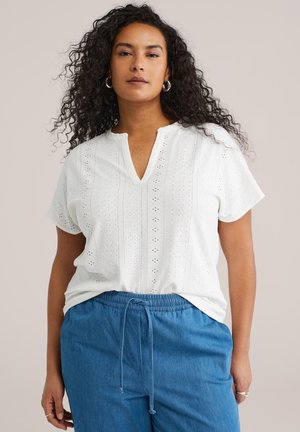 Femme aux cheveux bouclés portant un haut blanc à manches courtes ajourées et un pantalon bleu à cordon, sur fond uni.