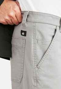 Pantalons en coton gris avec des détails de couture visibles, une poche arrière et un petit patch logo noir sur la taille. Main positionnée à la taille.