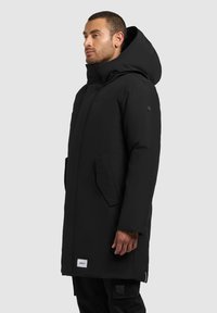 Čierna vonkajšia bunda s kapucňou, s elegantným dizajnom, so zapínaním na zips a dvoma bočnými vreckami. Vyrobená z hladkého, vodeodolného materiálu.