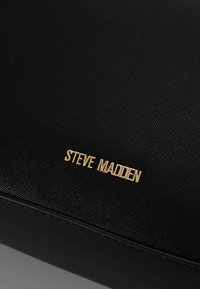 Sac à main noir texturé avec un accent logo doré "STEVE MADDEN". Le design est élégant avec une esthétique minimaliste.