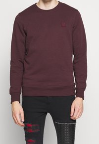 Bordowa bluza z okrągłym dekoltem, długimi rękawami i małą teksturowaną naszywką z logo na piersi. Wykonana z miękkiej, bawełnianej mieszanki materiałów.