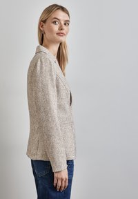 Beige strukturierte Blazer mit taillierter Passform, Ein-Knopf-Verschluss und zwei Fronttaschen, getragen über dunklen Denim-Jeans.