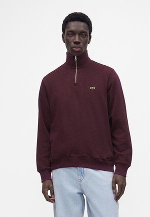 Mann trägt einen weinroten Sweatshirt mit Reißverschluss und hellblaue Jeans, steht vor einem einfarbigen weißen Hintergrund.