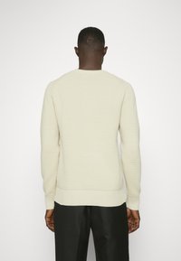 Cremfarbener Strickpullover mit einem strukturierten Muster, Rippbündchen und Saum. Getragen mit dunklen Hosen, die eine lässige Passform und ein entspanntes Design zeigen.