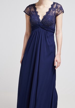 Kobieta w granatowej sukni wieczorowej z koronkowym gorsetem ozdobionym cekinami, z krótkimi rękawkami i przepływającą, sięgającą podłogi spódnicą.