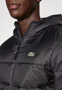 Black puffer jacket with a zip closure, featuring a green embroidered crocodile logo on the left chest and a hood. Smooth, shiny texture.