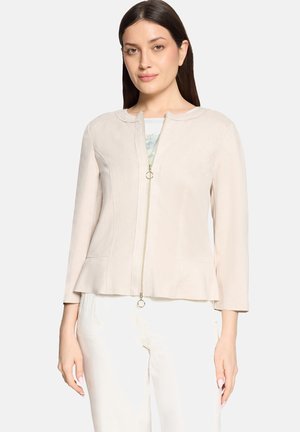 Kobieta o prostych ciemnych włosach, ubrana w jasnobeżową kurtkę na zamek, na wzorzystym topie i białych spodniach, stojąca na jednolitym tle.