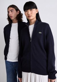Felpa con zip di colore navy in morbida maglia con polsini e orlo a costine. Presenta un piccolo logo di coccodrillo verde sul lato sinistro del petto.