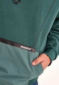 Sudadera verde con una sección inferior de un verde más oscuro. Presenta un logo negro y ribetes en los bolsillos, puños acanalados y una textura de tela suave.