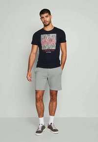 T-shirt de algodão azul-marinho com impressão gráfica e texto em contraste, combinada com calções cinza claro. O modelo usa meias brancas e ténis cinza.