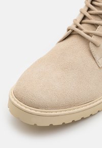 Zapato de ante beige con punta redondeada, diseño de cordones y superficie texturizada. Presenta una suela de goma de color claro.