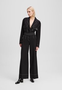 Black jumpsuit with lapel collar, front pockets, and belted waist. Features white stitching details, wide legs, and a smooth texture.