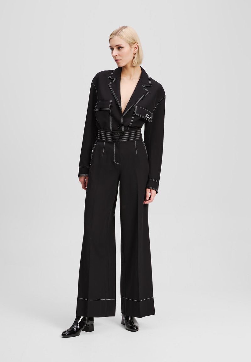 Black jumpsuit with lapel collar, front pockets, and belted waist. Features white stitching details, wide legs, and a smooth texture.