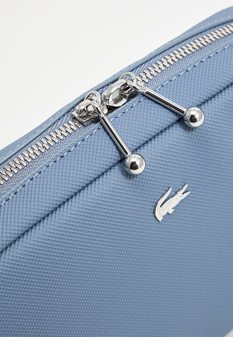 Pochette textile bleue avec surface texturée, dotée d'une fermeture éclair et de pièces métalliques en argent. Un petit logo de crocodile argenté est visible à l'avant.