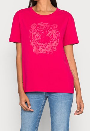 Femme portant un t-shirt rose vif avec un subtil graphisme de visage de tigre rose plus foncé, associé à un jean bleu clair.