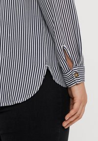 Chemise rayée bleu marine et blanche à manches longues, dotée d'un poignet boutonné et d'une fente sur le côté à l'ourlet, fabriquée dans un tissu léger.
