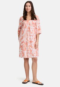 Rosa V-Ausschnitt-Kleid mit weißem Blattmuster, Dreiviertelärmeln, seitlichen Taschen und einer lockeren Silhouette. Getragen mit schwarzen Sandalen.